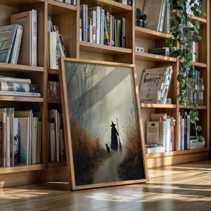 May include: A framed art print featuring a witch with a cat walking down a path in a misty forest. The artwork has a muted color palette with a wooden frame. The print is displayed on a wooden floor in front of a bookshelf.