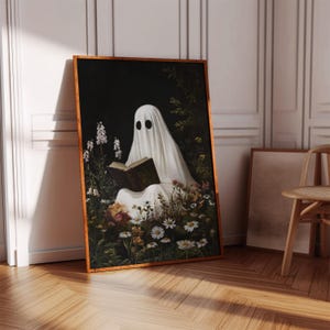 May include: A framed art print depicts a ghost reading a book while sitting in a flower garden. The ghost is draped in a white sheet with black eye holes. The artwork has a dark background and a wooden frame.