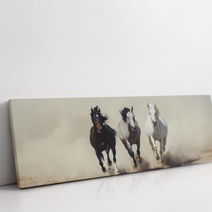 May include: A canvas print featuring three horses galloping through a dusty environment. The horses are a dark brown, gray, and white, with their manes and tails flowing in the wind. The artwork is rectangular and leans against a white wall.
