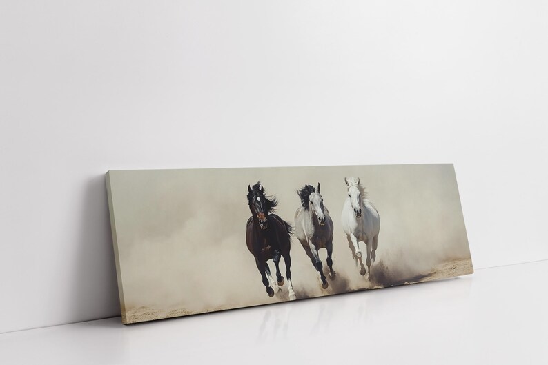 May include: A canvas print featuring three horses galloping through a dusty environment. The horses are a dark brown, gray, and white, with their manes and tails flowing in the wind. The artwork is rectangular and leans against a white wall.
