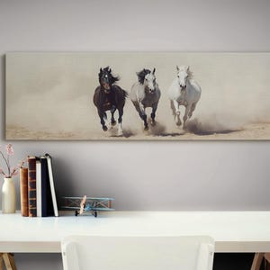 May include: A canvas print featuring three horses galloping across a dusty landscape. The horses are a dark brown, gray, and white, with their manes and tails flowing in the wind. The artwork is rectangular and mounted on a wall.