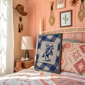 May include: A framed print depicting a cowboy on a rearing horse in blue ink on a cream background. The print is set against a blue and white checked fabric, framed in a dark grey frame. The scene is set in a bedroom with a western theme.