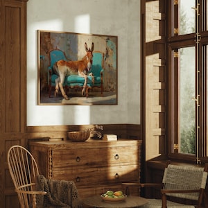 May include: A framed artwork shows a donkey reclining on a teal antique chair. The donkey has brown and white fur. The artwork is displayed in a room with wooden furniture, including a chest of drawers, chair, and window frame. The room is lit by natural light.