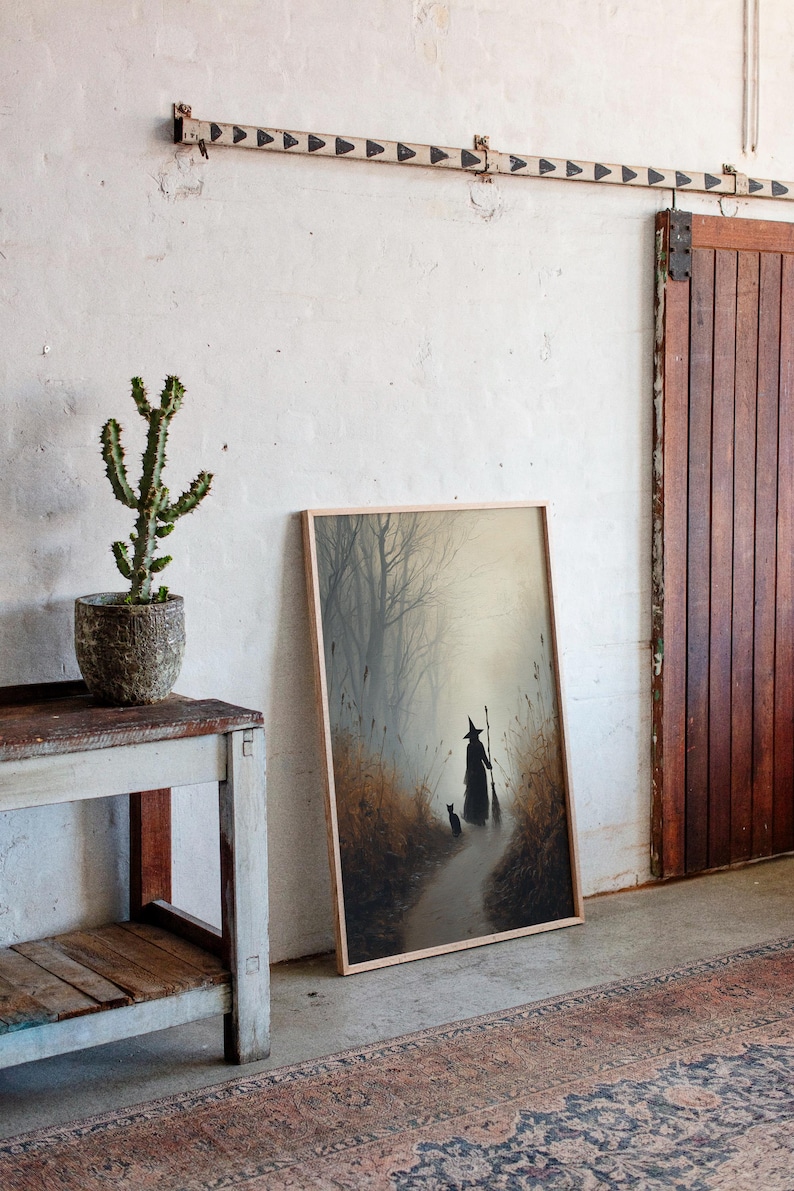 May include: A framed art print depicts a witch with a cat walking down a path in a misty forest. The artwork is in a light wood frame, leaning against a white wall. A cactus in a textured pot sits on a rustic wooden table.