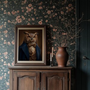 May include: A framed portrait of an owl dressed in a blue coat and bow tie, displayed on a wooden cabinet. The background features floral wallpaper and a terracotta vase with branches. Two lit candles add a warm glow.
