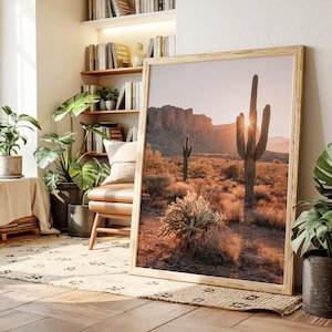 Può includere: Una stampa incorniciata di un paesaggio desertico con cactus e una catena montuosa al tramonto. La stampa ha una cornice in legno chiaro ed è appoggiata a un muro in una stanza con piante e libri.