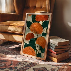 May include: A framed art print featuring orange chrysanthemum flowers with dark green leaves against a light background. The artwork is in a light wooden frame, leaning against a stack of books on a patterned rug.