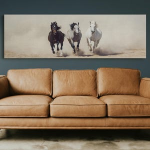 May include: A rectangular canvas print featuring three horses galloping across a sandy landscape. The horses are a dark brown, grey, and white. The print is mounted above a tan leather sofa with wooden legs.