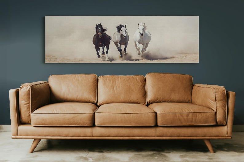 May include: A rectangular canvas print featuring three horses galloping across a sandy landscape. The horses are a dark brown, grey, and white. The print is mounted above a tan leather sofa with wooden legs.