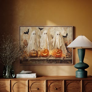 May include: A framed Halloween-themed painting featuring three white ghost figures and three carved pumpkins with glowing faces. Black bat silhouettes are scattered across the top. The artwork is displayed on a wooden cabinet with a green lamp.