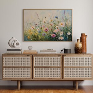May include: A framed floral painting with a wooden frame hangs above a wooden cabinet. The cabinet has woven rattan doors and is decorated with books, vases, and decorative objects. The painting features colorful wildflowers.