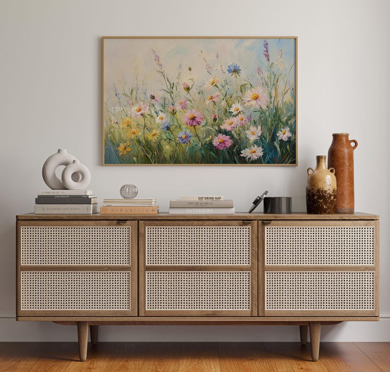 May include: A framed floral painting with a wooden frame hangs above a wooden cabinet. The cabinet has woven rattan doors and is decorated with books, vases, and decorative objects. The painting features colorful wildflowers.