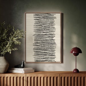 Könnte beinhalten: Abstrakter Kunstdruck in einem Holzrahmen. Das Kunstwerk zeigt einen beigefarbenen Hintergrund mit schwarzen gepunkteten Linien, die in vertikalen Reihen angeordnet sind. Der Rahmen hat eine hellbraune Farbe. Der Druck wird auf einem Holzschrank ausgestellt.