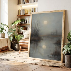 May include: A framed landscape painting depicts a serene, misty marsh scene. The artwork features a large, bright moon reflected in the calm water. Bare trees line the water's edge, and the overall color palette is muted with grays and browns.