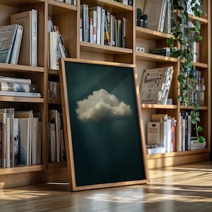 Puede incluir: Una impresión artística enmarcada que presenta una nube blanca y esponjosa sobre un fondo verde oscuro. La obra de arte se muestra en un marco de madera natural, apoyada contra una estantería llena de libros. La imagen evoca calma.