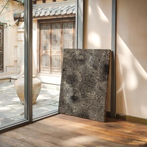 May include: A rectangular canvas print featuring a repeating pattern of black concentric circles on a cream background. The artwork is leaning against a wall near a window, with a wooden floor in the foreground.