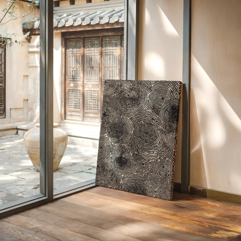 May include: A rectangular canvas print featuring a repeating pattern of black concentric circles on a cream background. The artwork is leaning against a wall near a window, with a wooden floor in the foreground.