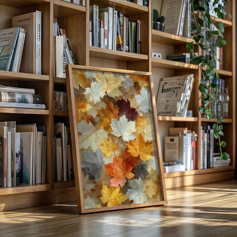 May include: A framed artwork depicting a colourful arrangement of autumn leaves in shades of yellow, orange, white, and burgundy. The artwork is displayed on a wooden floor, leaning against a bookshelf filled with books.