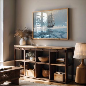 May include: A framed artwork depicting a tall ship sailing through icy waters, mounted above a wooden console table. The table features open shelves, drawers, and decorative items like vases, baskets, and books. A wooden lamp with a fabric shade is on the right.
