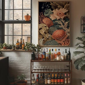 May include: A framed botanical print featuring coral and sea life in shades of peach, green, and cream. The artwork hangs above a bar cart filled with various bottles of liquor and glassware. A window with plants and bottles is in the background.