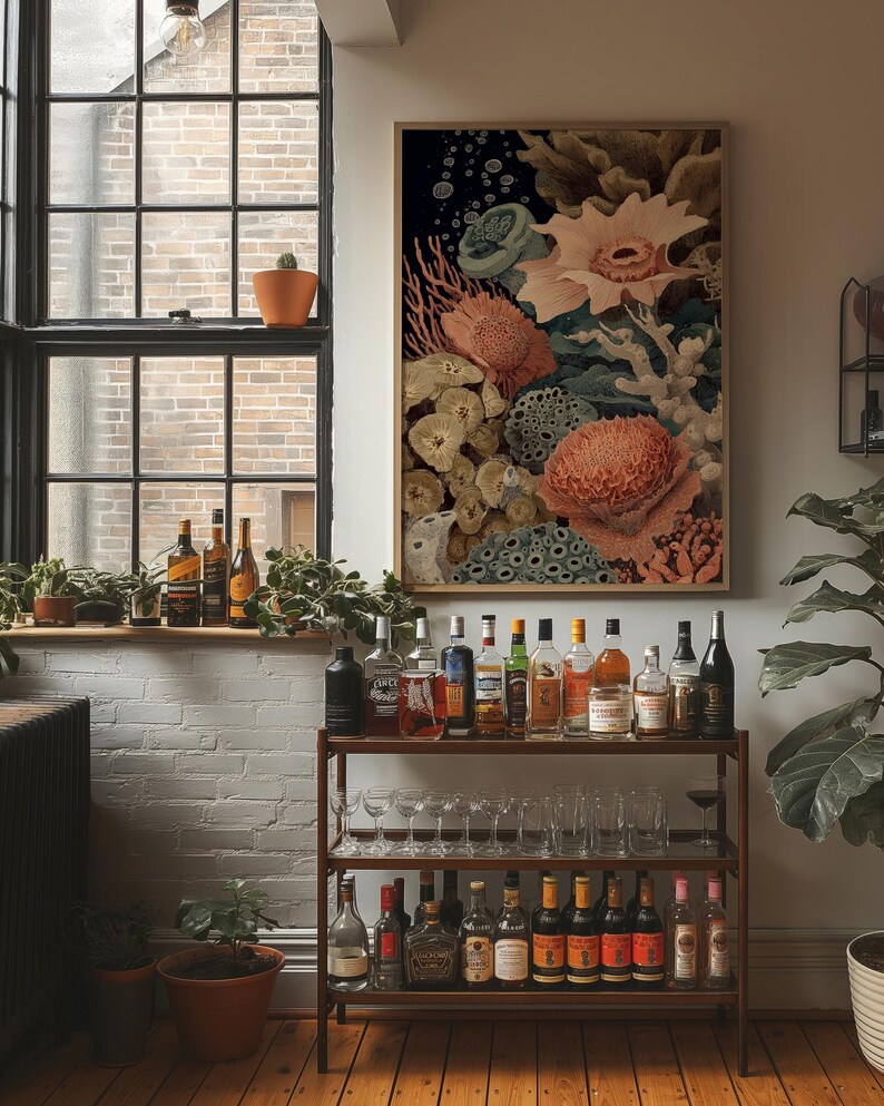 May include: A framed botanical print featuring coral and sea life in shades of peach, green, and cream. The artwork hangs above a bar cart filled with various bottles of liquor and glassware. A window with plants and bottles is in the background.
