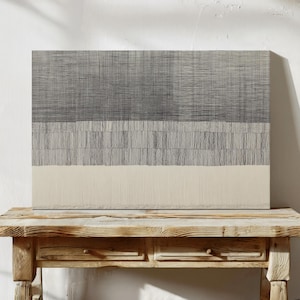 May include: Abstract canvas art with a layered design. The top section is a dark gray, the middle is a lighter gray, and the bottom is a cream color. The artwork is displayed on a rustic wooden table.