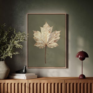 May include: A framed print featuring a close-up of a dried maple leaf against a muted green background. The leaf is off-white with brown spots and a long, thin, reddish-brown stem. The frame is a light brown color. The print is displayed above a wooden cabinet.