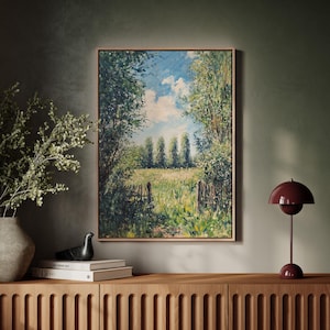 May include: A framed landscape painting depicts a sunny field with a wooden fence, trees, and a blue sky with clouds. The artwork is displayed above a wooden cabinet, next to a vase with greenery and a burgundy table lamp.