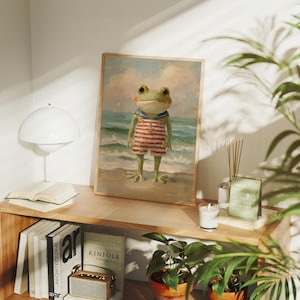 May include: A framed art print depicts a frog dressed as a sailor standing on a beach. The frog wears a red and white striped outfit with a blue collar. The background shows a seascape with a sailboat under a cloudy sky.