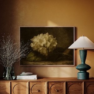 Könnte beinhalten: Ein gerahmter Kunstdruck mit weißen Hortensienblüten und grünen Blättern. Eine türkisfarbene Lampe mit weißem Lampenschirm steht auf einem Holzschrank. Eine Glasvase mit getrockneten Zweigen befindet sich links.