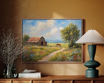 Puede incluir: Pintura enmarcada de un paisaje rural con un granero y un caballo en un campo de flores silvestres. Un camino de tierra conduce al granero bajo un cielo azul con nubes. La pintura se exhibe sobre un mueble de madera. El marco es de madera clara.