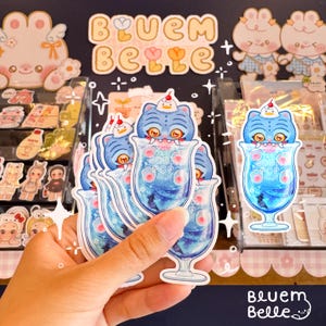 May include: A collection of stickers featuring a blue tiger character in a soda pop glass. The stickers have a blue and white color scheme with the words "Soda Pop" on the glass. The stickers are held in a hand.