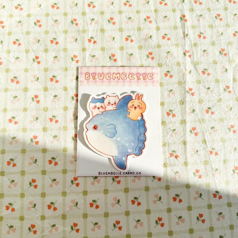 May include: A sticker featuring a whimsical illustration of a blue fish with three cartoon characters riding on its back. The sticker has the text "BYUEMBELLE" at the top and "BLUEMBELLE.CARRD.CO" at the bottom. The sticker is on a floral patterned background.