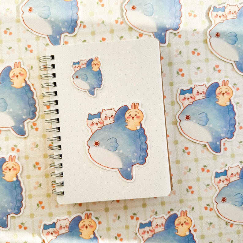 May include: A spiral-bound notebook with a white cover is surrounded by stickers. The stickers feature a blue fish with a smiling face, and a group of small, cute animals on top. The stickers are scattered on a floral patterned surface.