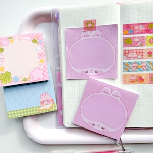 May include: A collection of stationery items, including pink pens, sticky notes, and decorative tape. The sticky notes feature a cute bunny design and the text "BRAIN DUMP". The items are arranged on a white tray and inside a planner.