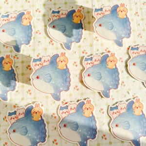 May include: A collection of stickers featuring a blue fish with a smiling face, a small fin, and a decorative edge. Each fish has a small orange bunny and a group of white and gray bears on top. The stickers are on a floral background.