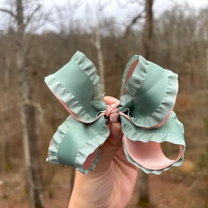 May include: A large, handmade hair bow with a layered design. The bow features a soft, sage green exterior with a delicate ruffled edge, and a light pink interior. The bow is tied in the center with a matching pink ribbon.