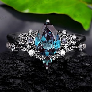 May include: A two-piece ring set featuring a pear-shaped, color-changing alexandrite gemstone. The ring is set in a dark metal with floral and leaf accents, and small white gemstones. The band has a matching design.
