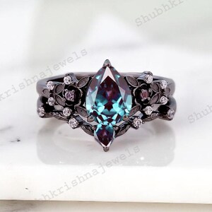 May include: A black metal ring set with a marquise-cut, teal-colored gemstone. The ring is adorned with floral and small diamond accents. The band has a matching floral design. The ring is set on a white surface.
