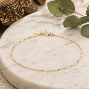 Peut inclure: Fin bracelet en or avec une chaîne fine et un fermoir mousqueton. Le bracelet est présenté sur une surface en marbre blanc, avec un élément décoratif doré en arrière-plan. Un bijou simple et élégant.