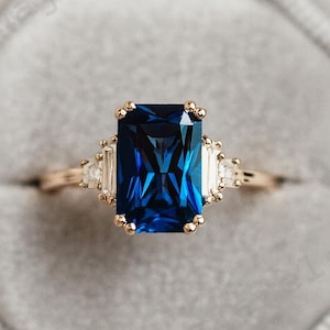 May include: A gold ring featuring a large, rectangular, deep blue gemstone. The ring is flanked by smaller, clear gemstones. The ring is presented in a grey velvet jewellery box.
