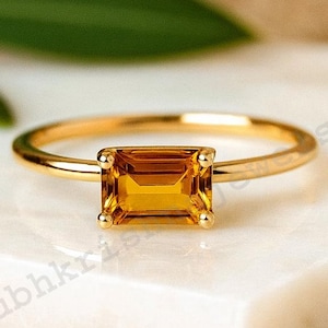 May include: A gold ring featuring a rectangular, amber-hued gemstone. The ring is set against a white surface with a blurred backdrop of green foliage and a neutral tone. The ring's band is slender and polished.