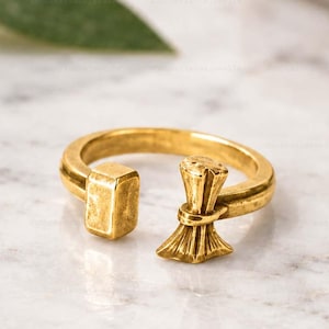 Op de afbeelding: Een goudkleurige open ring met een getextureerd rechthoekig blok aan het ene uiteinde en een uitlopend, waaiervormig element met verticale ribbels aan het andere uiteinde. Het metaal heeft een licht matte, geborstelde afwerking en de ring is rond.