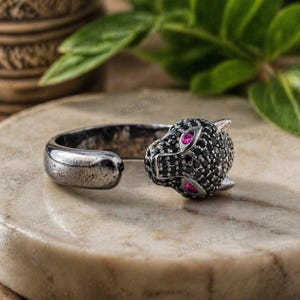 Könnte beinhalten: Ein silberfarbener Ring mit einem Pantherkopf-Design. Der Pantherkopf ist mit schwarzen Steinen besetzt und hat rosa Edelsteinaugen. Der Ring hat ein offenes Design, mit dem Pantherkopf an einem Ende und einem abgerundeten Band am anderen.