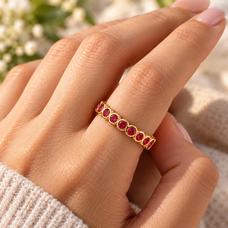 May include: A gold band ring featuring a continuous row of round, bezel-set vibrant red stones. The ring has a polished metallic finish and a uniform design, with each stone securely encased in a circular gold setting along the entire band.