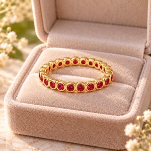 May include: A gold eternity band featuring a continuous row of round, vibrant red gemstones set in individual circular bezels. The ring is displayed inside a soft, light beige velvet jewellery box, highlighting the polished gold metal.