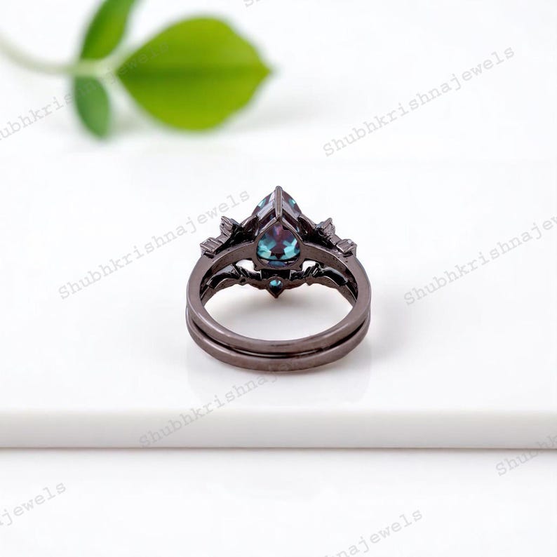 May include: A dark gray ring set with a pear-shaped, teal-colored gemstone. The ring has a matching band and intricate detailing around the main stone. The jewelry is set against a white background.