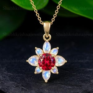 Op de afbeelding: Een gouden ketting met een hanger met een centrale rode edelsteen omringd door witte en blauwe druppelvormige stenen, die een bloemmotief vormen. De ketting en hanger zijn goudkleurig, tegen een donkere achtergrond.