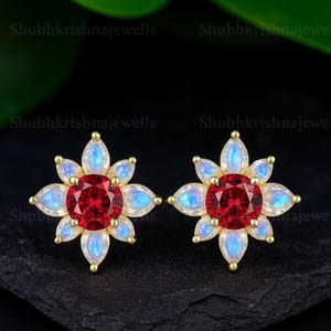 May include: A pair of gold-coloured stud earrings. Each earring has a central, round, red gemstone surrounded by six light blue and white petal-shaped stones. The earrings are set against a dark background.