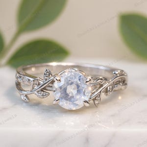 May include: A silver ring with a large, round, clear gemstone. The band has a leaf and vine design with small accent stones. The ring is set against a marble surface.